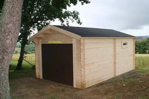 Garage en madriers, porte brune, lisière de bois